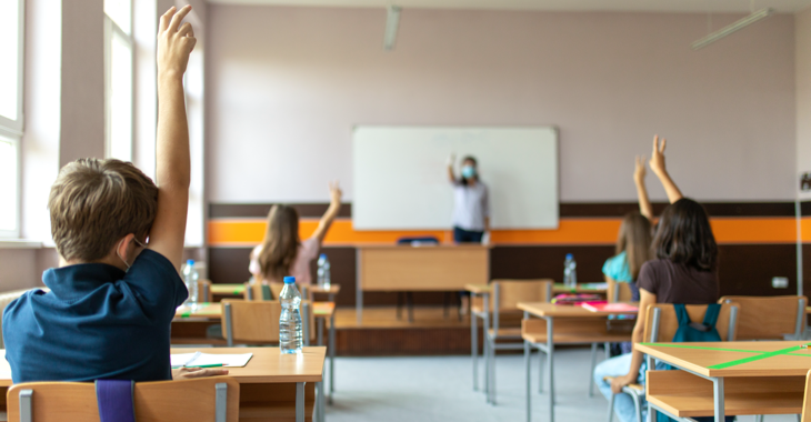 Schüler:innen mit Masken in einem Klassenzimmer melden sich, im Hintergrund eine Lehrerin mit Maske vor der Tafel © iStock/miljko Schüler:innen mit Masken in einem Klassenzimmer melden sich, im Hintergrund eine Lehrerin mit Maske vor der Tafel © iStock/miljko