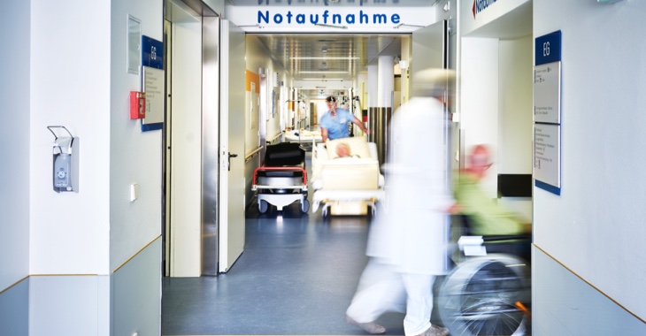 Krankenpfleger und Arzt schieben Rollstuhl und Fahrtrage in einem Krankenhausflur, darüber ein Schild mit Aufschrift Notaufnahme © iStocj/upixa