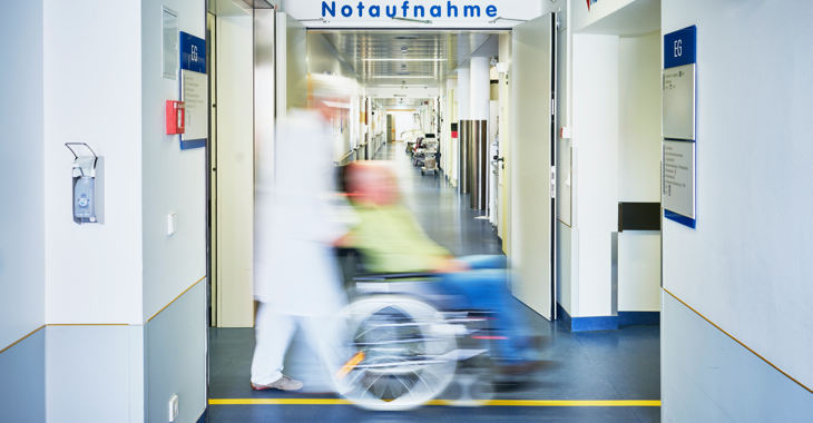 Ein Pfleger schiebt in einem Krankenhaus einen Patienten im Rollstuhl durch einen Flur mit einem Schild Notaufnahme an der Wand © iStock/upixa