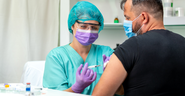 Krankenschwester mit Schutzbrille und Maske verabreicht einem Mann mit Maske eine Injektion @ iStock/zoranm