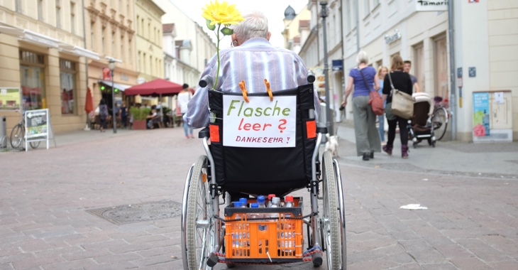 Ein alter Mann im Rollstuhl mit einem Zettel »Flasche leer? Danke sehr!« sammelt in einer Fußgängerzone leere Flaschen © dpa/Kay Nietfeld