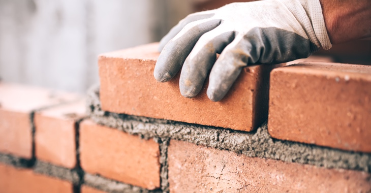 Eine Hand mit Handschuh hält einen Ziegelstein auf einer unvollständigen Wand mit frischem Mörtel © iStock/Bogdanhoda