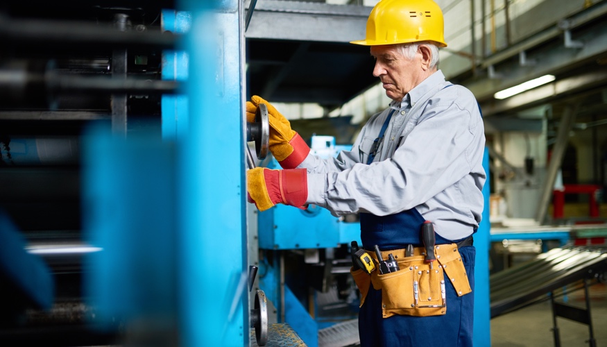 Ein älterer Arbeiter mit gelbem Schutzhelm schraubt an einer blauen Maschinen © iStock/SeventyFour