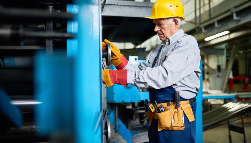 Ein älterer Arbeiter mit gelbem Schutzhelm schraubt an einer blauen Maschinen © iStock/SeventyFour Ein älterer Arbeiter mit gelbem Schutzhelm schraubt an einer blauen Maschinen © iStock/SeventyFour
