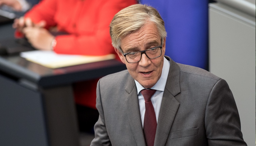 Dietmar Bartsch am Rednerpult des Bundestag, dahinter verschwommen Kanzlerin Merkel in roter Jacke © Soeren Stache/dpa