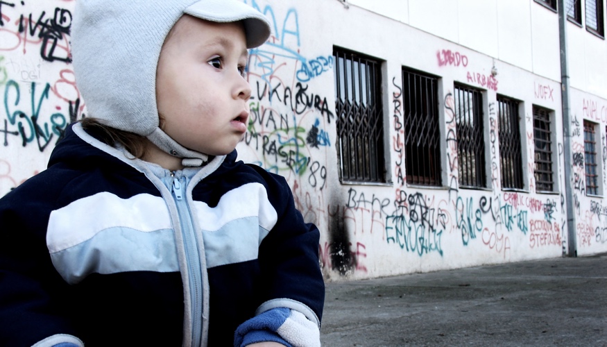 Ein Kind vor einem Haus mit Graffiti an der Wand und Gittern vor den Fenstern © iStock/brunette